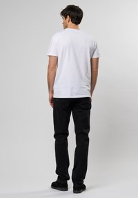 Tricou alb din bumbac cu mâneci scurte, combinat cu pantaloni din denim negri și pantofi negri, văzut din spate pe un fundal gri.