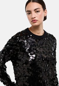 Pull noir à sequins avec encolure ronde et surface texturée, orné de sequins circulaires qui reflètent la lumière.
