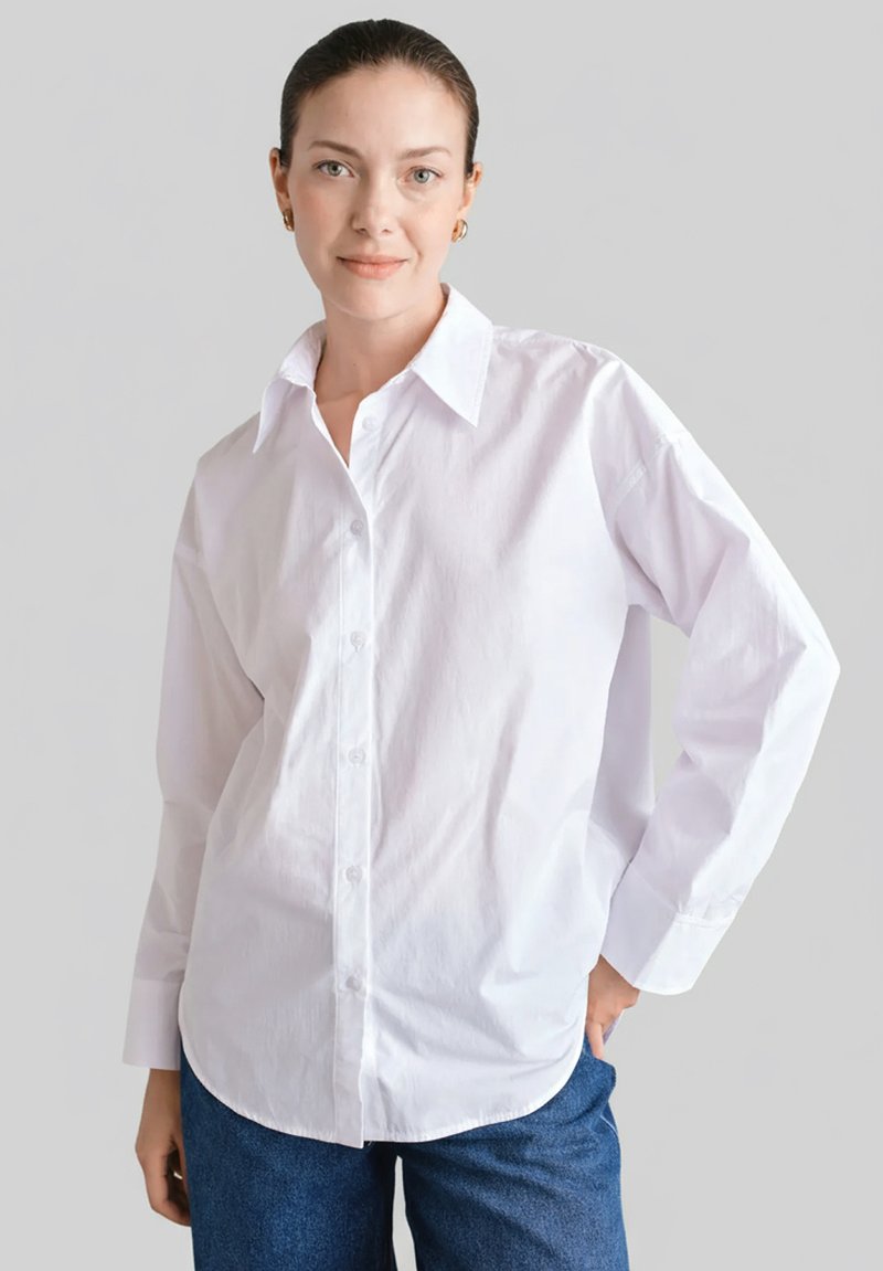 Biała koszula na guziki z ostrym kołnierzykiem, długimi rękawami i lekko luźnym krojem. Noszona z niebieskimi dżinsowymi spodniami. Gładka bawełniana tkanina.