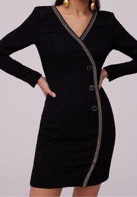 Robe noire texturée à manches longues avec fermeture diagonale à boutons et bordure décorative, portée avec de grandes boucles d'oreilles rondes dorées.