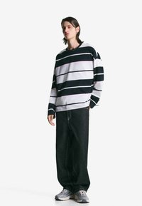 Ung person iført en sort-hvid stribet langærmet skjorte, løse sorte jeans og grå sneakers, stående foran en hvid baggrund.