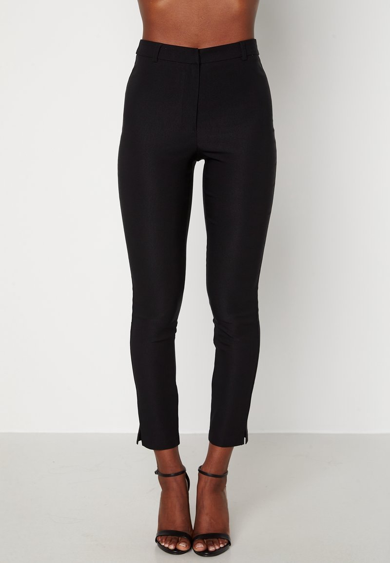 Pantalon skinny noir taille haute avec une texture lisse, coupe ajustée et fentes latérales à l'ourlet. Présente une ceinture classique et aucun élément métallique visible.