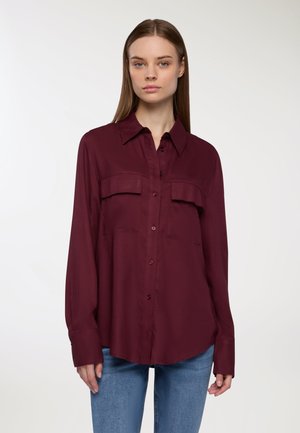 Camicia bordeaux con bottoni in tessuto liscio, dotata di due tasche sul petto, maniche lunghe e orlo leggermente rotondo. Abbinata a jeans di denim.
