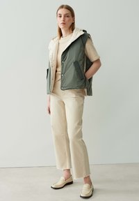 Padded Weste in Grün und Creme, über einem beigen Pullover getragen, kombiniert mit off-white weit geschnittenen Hosen und beigen Slippern, vor einem hellen Hintergrund.