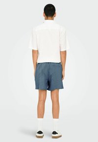 White short-sleeve shirt, textured blue shorts, white socks, and black and beige shoes. Viewed from the back against a neutral background.