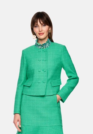 Woman wearing a teal textured double-breasted jacket and matching skirt, with a patterned ruffle collar shirt and large gold earrings.