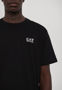 Svart bomulls t-skjorte med en liten hvit "EA7 Emporio Armani"-logo på brystet og korte ermer. Glatt tekstur med en klassisk passform.