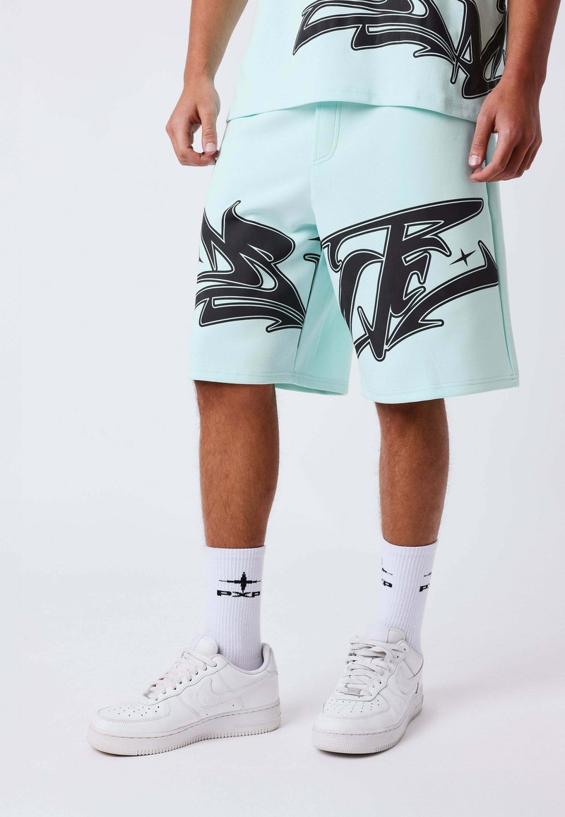 Personne portant un short bleu clair avec un imprimé style graffiti noir, des chaussettes hautes blanches avec logos, et des baskets blanches, se tenant sur un fond uni.