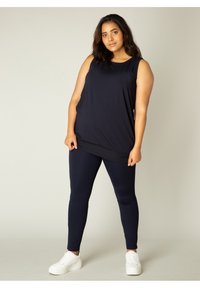 Marineblauwe tanktop met een ontspannen pasvorm, gecombineerd met smal gesneden marineblauwe leggings. Beide zijn gemaakt van een zachte stof. Model draagt witte sneakers.