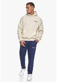 Beige hættetrøje med forbagslomme og "Dropsize"-logo; parret med navy sweatpants, som også har "Dropsize"-logoet. Hvide sneakers.