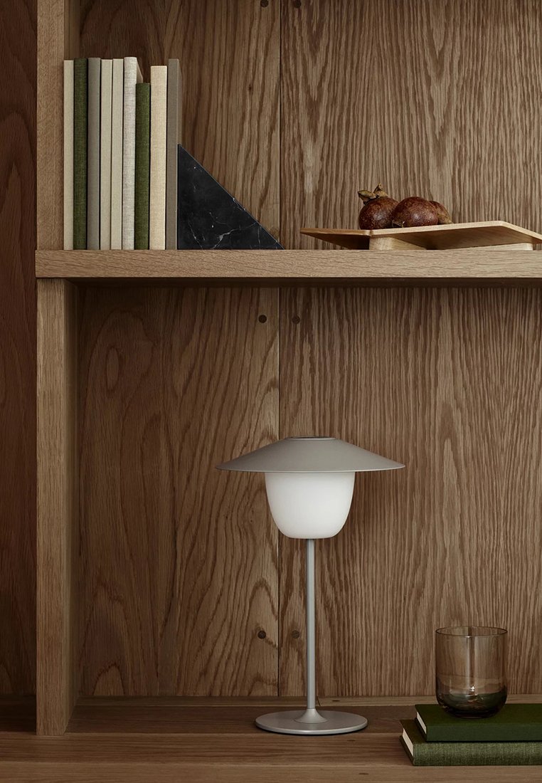 Holzregal mit Büchern und einem schwarzen Buchstütze, ein Holzteller mit Obst, eine moderne Tischlampe, ein Glas und zwei übereinander gestapelte Bücher.
