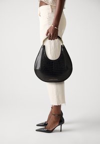 Woman in cream cropped pants and black heels holding black crocodile-textured handbag with gold handles at side.