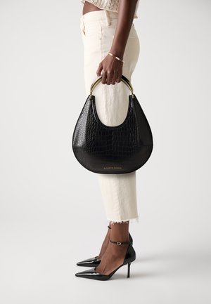 Woman in cream cropped pants and black heels holding black crocodile-textured handbag with gold handles at side.