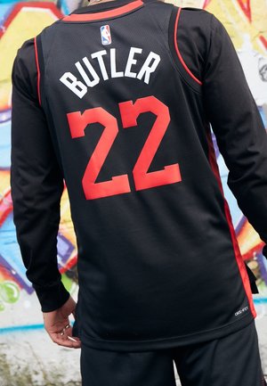Person wearing a black basketball jersey with red number 22 and name Butler, standing in front of a colorful graffiti wall.