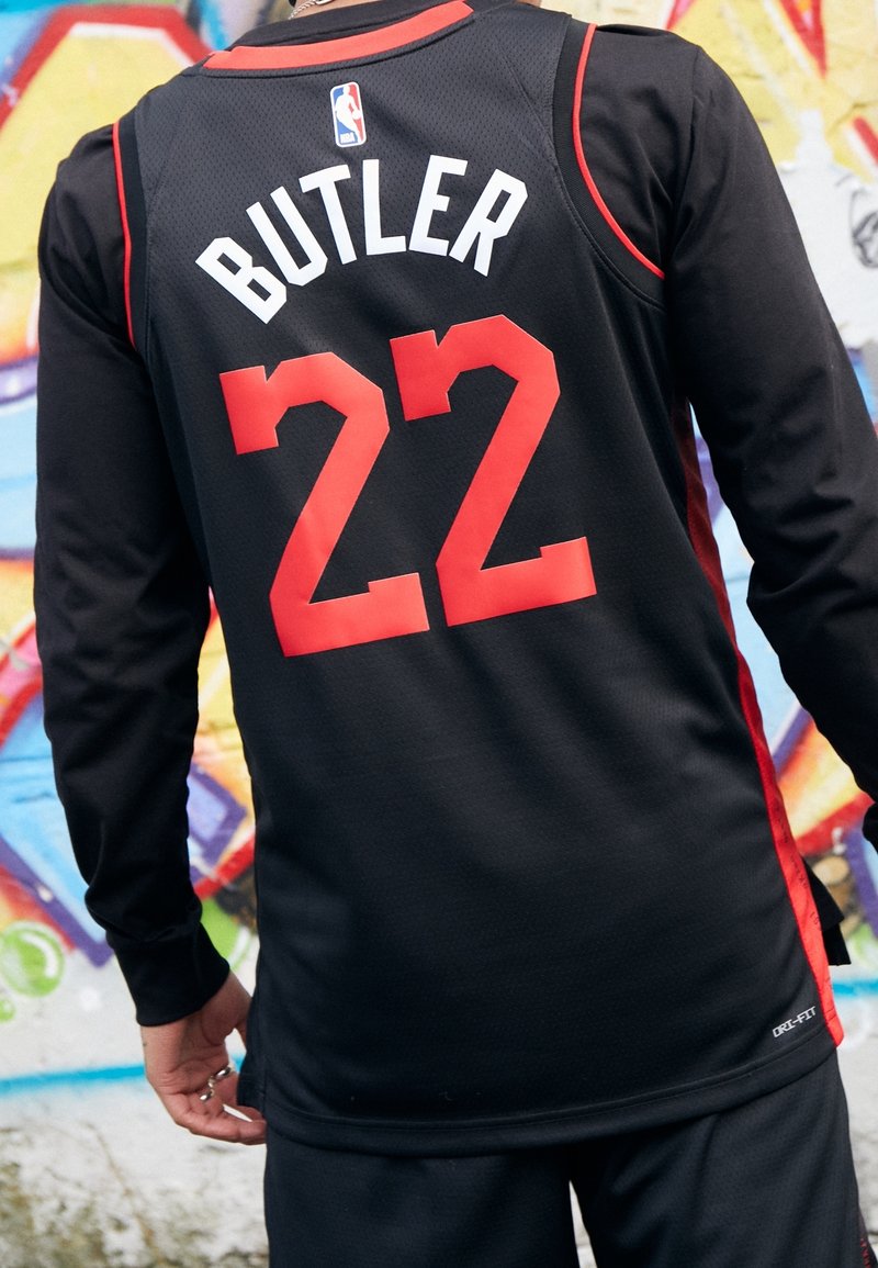 Personne portant un maillot de basket noir avec le numéro rouge 22 et le nom Butler, debout devant un mur de graffiti coloré.