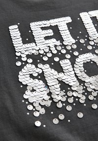 Gray fabric t-shirt featuring the text "LET IT SHINE" in large, glittering silver sequins, scattered decoratively across the material.