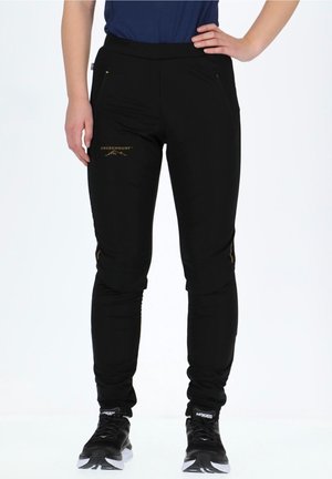 Personne portant un pantalon de sport noir de la marque Swedemount et des chaussures de course Hoka noires et blanches, debout avec une main sur la hanche.