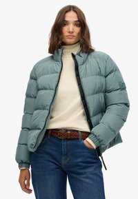 Puff jakke i dæmpet teal, med lynlås foran, høj krave og elastiske manchetter, parret med en beige sweater og blå denim jeans.