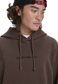 Hnedá mikina s kapucňou a šnúrkou na stiahnutie, s čiernym vyšitým logom "Quiksilver". Jednoduchý dizajn a mäkká textúra látky. Voľný strih.
