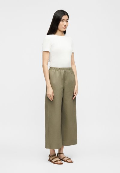 Ženska stoji postrani, oblečena v belo majico s kratkimi rokavi, ohlapne olivne široke hlače z elastičnim pasom in rjave ravne sandale na enobarvnem ozadju.