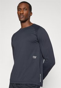 Camisola atlética de manga longa em azul marinho escuro, com decote redondo, mangas raglan e uma etiqueta com o logótipo no lado inferior esquerdo. Textura de tecido suave.