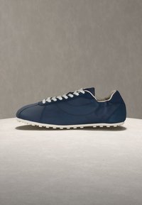 Αθλητικό παπούτσι από δέρμα σε ναυτικό μπλε με λευκά κορδόνια και σόλα, τοποθετημένο σε μια ανοιχτή, στρογγυλή επιφάνεια με φόντο απλό γκρι.