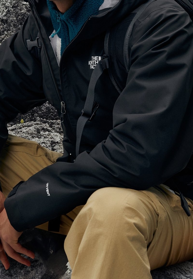 Veste imperméable noire avec poignets ajustables, sangles grises et un logo sur la poitrine. Portée sur une polaire bleue et associée à un pantalon beige.