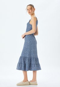 Blauwe bloemen midi-jurk met spaghetti bandjes, gerimpelde zoom en lichtgewicht stof. Beschikt over een aansluitend lijfje en een uitlopende rok. Draagt met beige instappers.