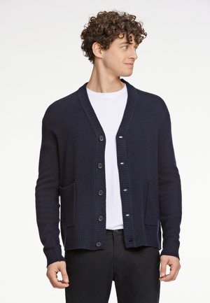 Granatowy kardigan, dzianinowa struktura, pięć guzików, dwie przednie kieszenie, ściągacze przy mankietach, noszony na białej koszulce, casualowy design.