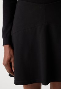Jupe patineuse noire en tissu doux et flexible. Présente une taille ajustée, une longueur en dessous du genou et de subtils détails de couture à l'ourlet.