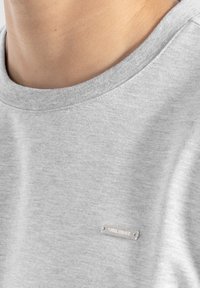 T-shirt grigio in cotone con scollatura rotonda e una piccola etichetta metallica nella zona sinistra del petto. Texture morbida con una finitura liscia.