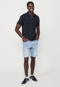 Námornícky polo tričko s malým bodkovaným vzorom, skombinované s svetlými riflovými šortkami. Model má na sebe tmavé tenisky a stojí so založenými rukami v vreckách.
