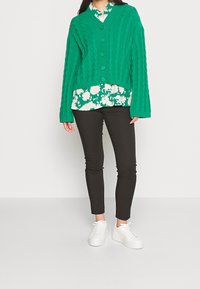 Cardigan vert en maille torsadée avec cinq boutons, porté sur un chemisier blanc à fleurs. Pantalon noir ajusté et baskets blanches complètent la tenue.