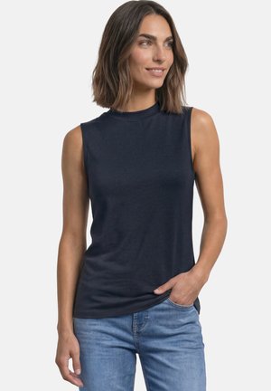 Donna con capelli castani a caschetto che indossa una canotta nera senza maniche e jeans blu, sorridente e che guarda di lato con una mano in tasca.