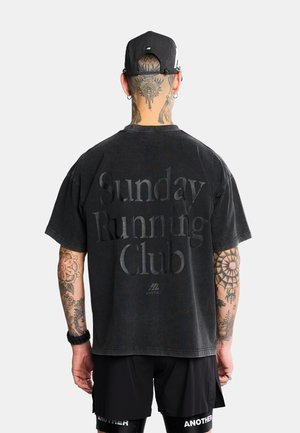 Uomo con tatuaggi che indossa un cappellino nero, una T-shirt nera oversize con il testo "Sunday Running Club" e pantaloncini neri, rivolto di spalle alla fotocamera.