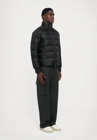 Mand iført en sort quilted jakke, mørke cargo bukser og hvide sneakers står foran en ensfarvet hvid baggrund.