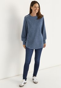 Lächelnde Frau trägt einen blauen, langärmeligen Strickpullover mit Metallknöpfen an der Schulter, dunkle Jeans und weiße Sneaker, während sie vor einer weißen Wand steht.