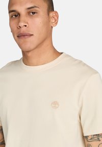 Béžové tričko s krátkým rukávem z měkké tkaniny, které na levé straně hrudi zdobí malé vyšití loga stromu. Design s kruhovým výstřihem, minimalistický styl.