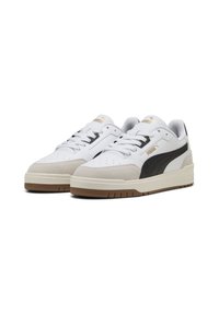 Baskets de sport blanches et noires avec des accents en daim et une semelle en caoutchouc texturé. Elles présentent des perforations sur le bout et un logo doré sur la languette.