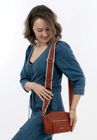 Brown textured leather crossbody bag with a zippered front pocket and patterned adjustable strap worn over a blue denim outfit.