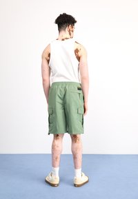 Pantalones cortos de carga verdes, fabricados con material ligero, con múltiples bolsillos y cinturilla elástica, combinados con una camiseta sin mangas blanca y zapatillas beige.