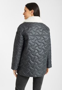 Grijze gewatteerde jas met een witte faux fur kraag, voorzien van een getextureerd spiraalpatroon en een losvallend ontwerp. De achterkant laat de schoudernaden zien.