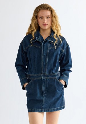 Giovane donna con lunghi capelli biondi e ricci, indossa un vestito giacca di jeans blu scuro con le mani nelle tasche, rivolta in avanti su sfondo semplice.