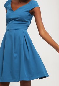 Eine Frau trägt ein figurbetontes, blaues, ärmelloses Kleid mit einem schwingenden Rock vor einem schlichten, hellen Hintergrund.