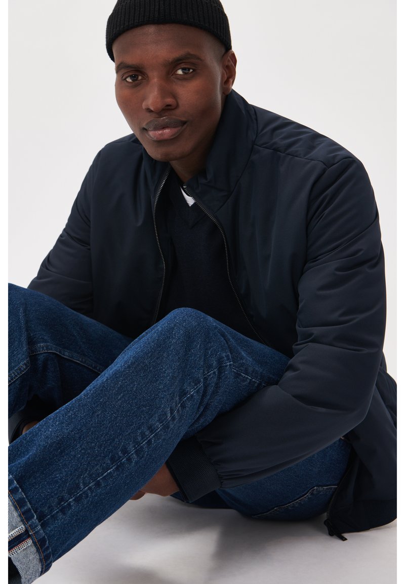 Jūrinis pūkuotas švarkas su pakeltu apykakle, derinamas su tamsiai mėlynomis džinsinėmis kelnėmis. Švarkas turi užtrauktuką ir raštuotus rankogalius.