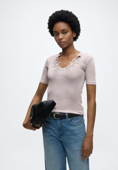 Νεαρή γυναίκα με κοντά σγουρά μαλλιά φοράει ανοιχτό ροζ τοπ με δαντέλα, μπλε τζιν, μαύρη ζώνη και κρατά μια μαύρη δερμάτινη τσάντα με απλό φόντο.