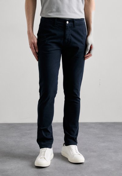 Pantalones ajustados de color azul marino, hechos de tela gruesa, con cremallera frontal, cierre de botón y bolsillos laterales. Combinados con zapatillas blancas.