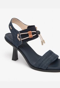 Denim højhælede sandal med en bred rem, kontrastfarvet navy indersål og en justerbar ankelrem med en tan læderdetalje og dekorativ hardware.