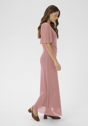 Kobieta z długimi brązowymi włosami, ubrana w długą sukienkę w kolorze przybrudzonego różu oraz brązowe, niskie, plecione buty na obcasie, stojąca bokiem na białym tle.