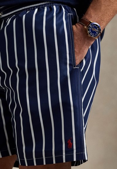 Nautičko plave kupaće gaće s bijelim okomitim prugama, elastičnim pojasom, bočnim džepovima i crvenim izvezenim logotipom. Glatka tekstura tkanine.